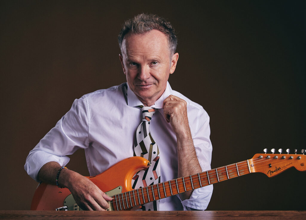 Ein Herr in rosa Hemd schaut in die Kamera und hält seine E-Gitarre in der Hand