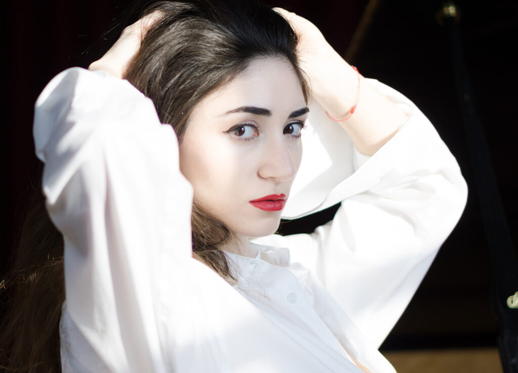 Porträt einer jungen Frau mit roten Lippen und weißer Bluse, die sich mit beiden Händen durch das Haar fährt. Das Licht fällt dramatisch auf ihr Gesicht.