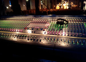 ein großes Tonmischpult mit vielen leuchtenden Lämpchen in einer Indoor Bühnen Umgebung