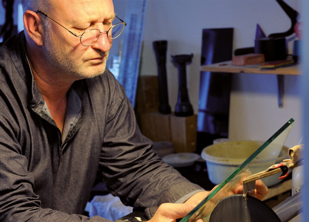 Ein älterer Mann mit Brille arbeitet konzentriert an einem Stück Glas in einer Werkstattumgebung.