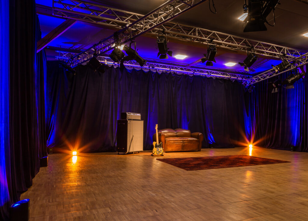 Professionelles Aufnahmestudio mit Sofa, Verstärker und E-Gitarre auf einer Bühne, beleuchtet von blau-orangem Bühnenlicht